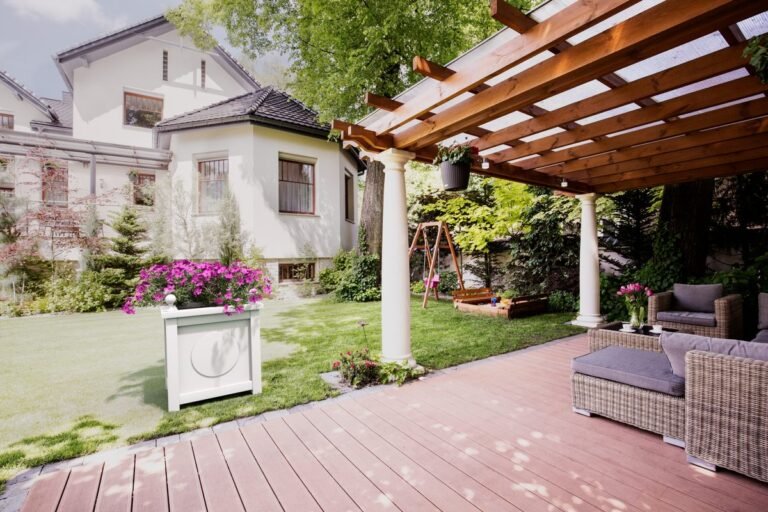 Ein weißer dekorativer Pflanzkübel mit blühenden Blumen steht auf einem Rasen neben einer Terrasse mit Pergola vor einem Haus mit Garten