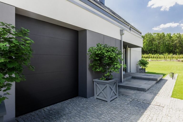 Ein grauer quadratischer Pflanzkübel mit einem kleinen Baum steht vor einem modernen Haus neben einer Garage auf einer gepflasterten Einfahrt