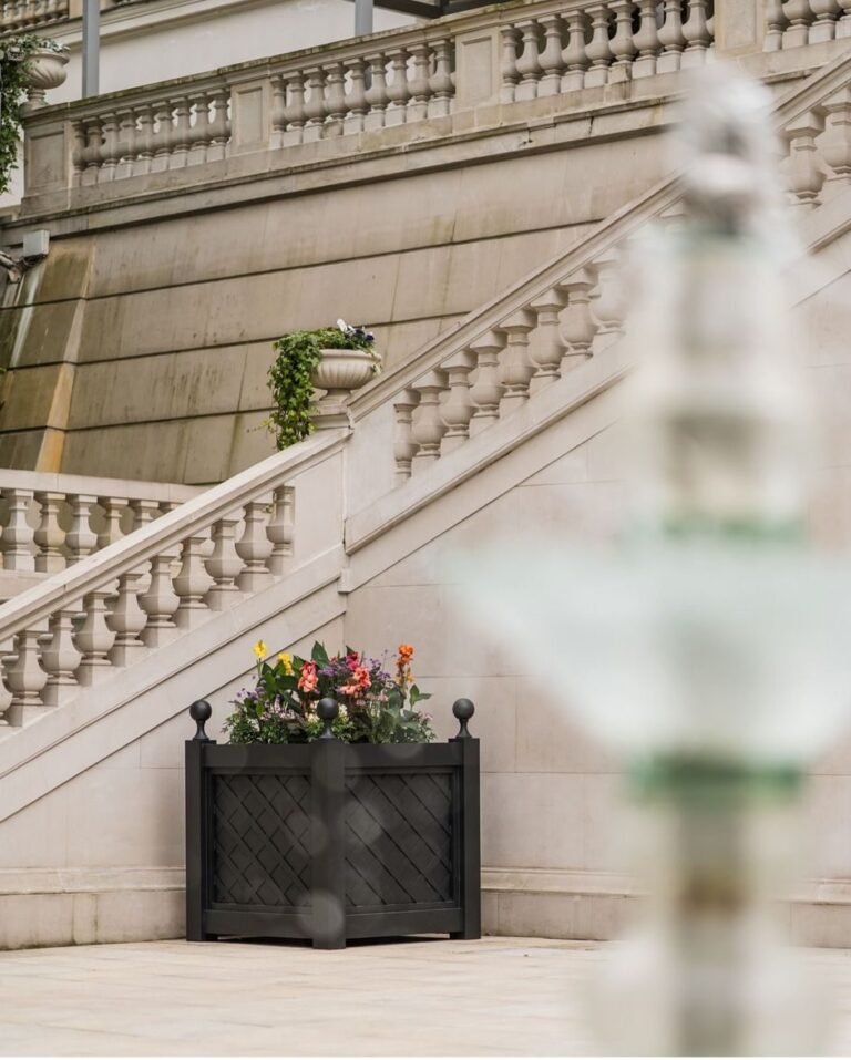 Ein schwarzer dekorativer Pflanzkübel mit Blumen steht vor einer klassischen Steintreppe mit Balustrade an einem eleganten Gebäude