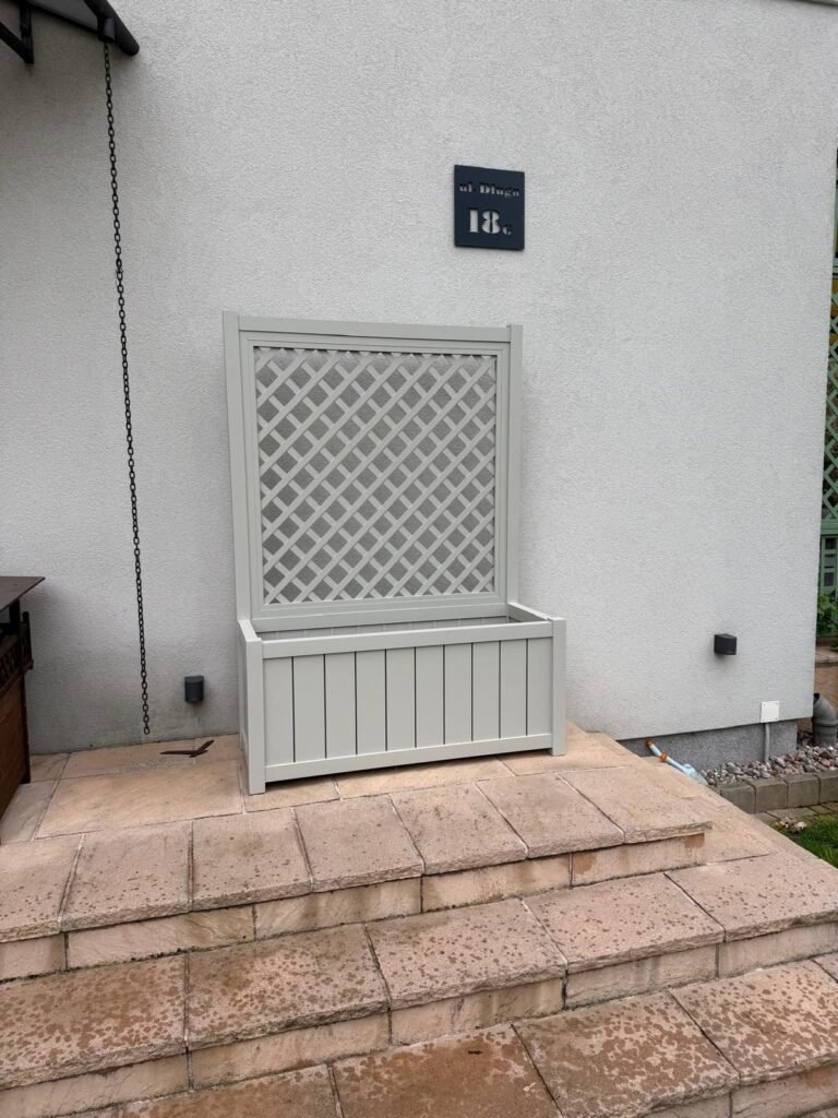 Ein hellgrauer Pflanzkasten mit integriertem Rankgitter steht auf einer Terrasse vor einer Hauswand neben einer Treppe