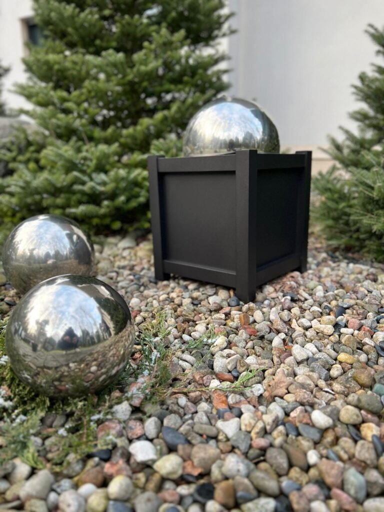 Ein schwarzer quadratischer Pflanzkübel steht in einem Steingarten neben dekorativen glänzenden Metallkugeln, umgeben von kleinen Nadelbäumen und Sträuchern