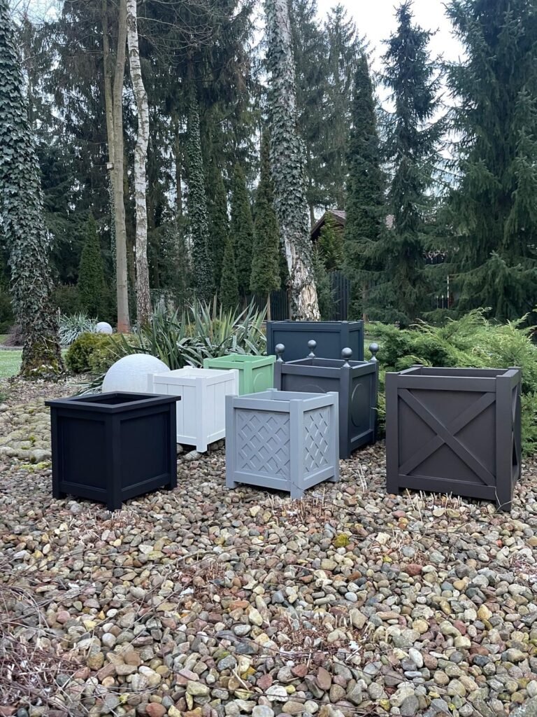 Mehrere quadratische Pflanzkübel in verschiedenen Farben stehen auf einem Steingarten in einem bewaldeten Garten mit hohen Bäumen und Sträuchern