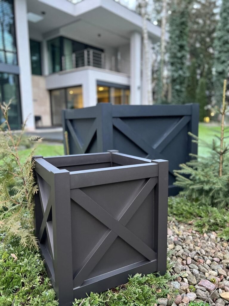 Zwei schwarze quadratische Pflanzkübel stehen in einem modernen Garten mit Steinen und kleinen Nadelpflanzen vor einem zeitgenössischen Haus mit großen Fenstern und Balkon
