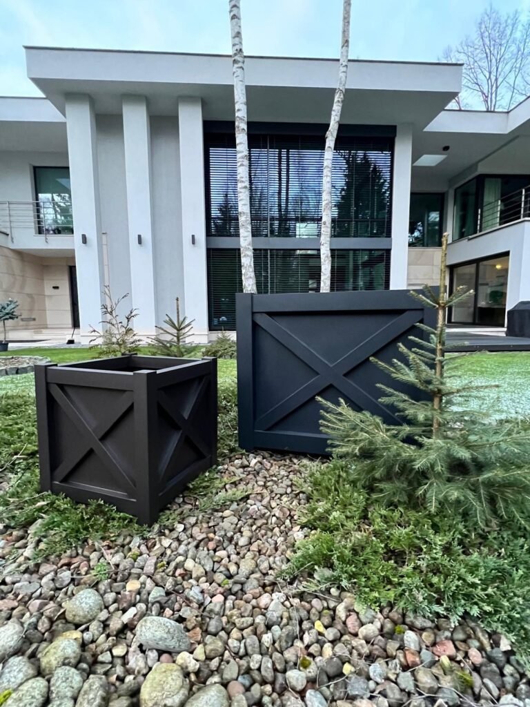 Zwei schwarze quadratische Pflanzkübel stehen in einem Steingarten vor einem modernen Haus mit großen Fenstern, umgeben von kleinen Nadelbäumen und Birken