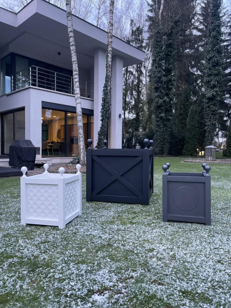 Drei quadratische Pflanzkübel in Weiß, Schwarz und Grau stehen auf einem leicht verschneiten Rasen vor einem modernen Haus mit großen Fenstern und einem Balkon, umgeben von hohen Bäumen