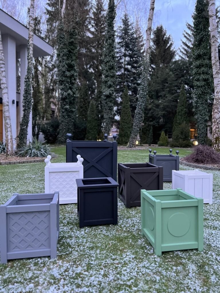 Mehrere quadratische Pflanzkübel in verschiedenen Farben stehen auf einem Rasen in einem modernen Garten mit hohen Bäumen neben einem zeitgenössischen Haus