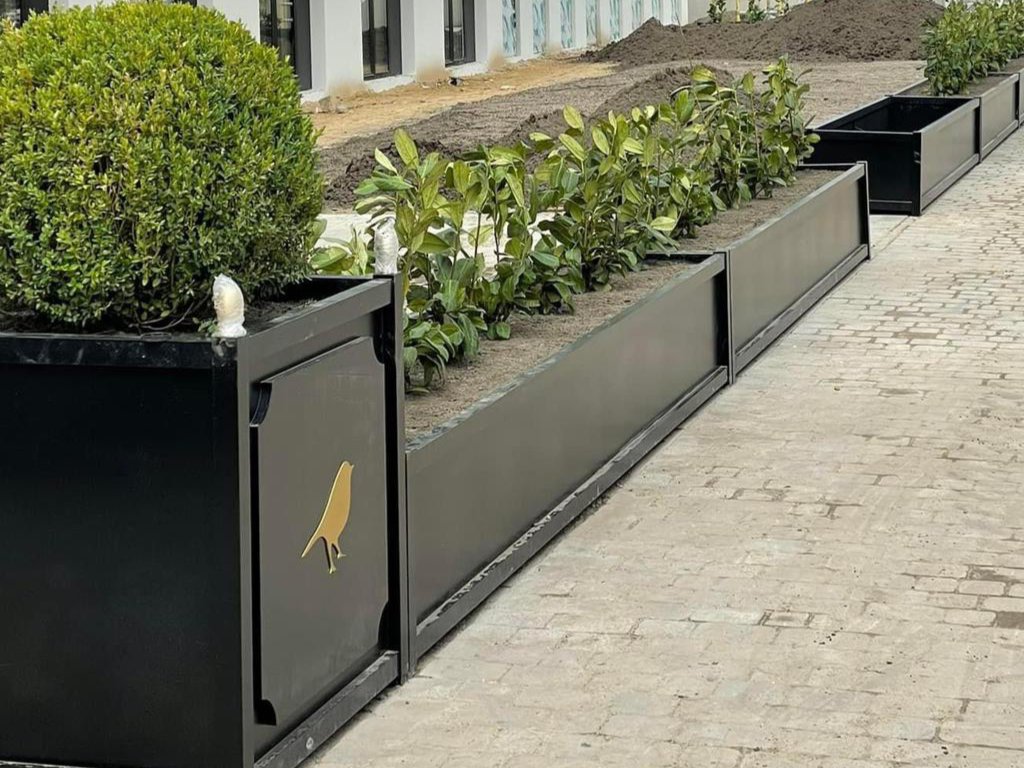 Exklusive Pflanzkübel Outdoor aus Aluminium entlang eines gepflasterten Weges vor einem modernen Gebäude, bepflanzt mit grünen Sträuchern und einem runden Buchsbaum.